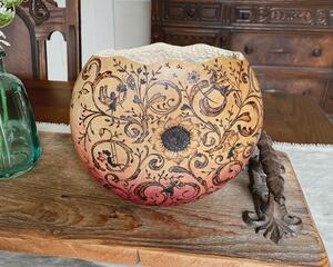 A round vase sits on a dining table. It is a natural gourd with whimsical fairies and flourish designs burned onto the gourd shell. 